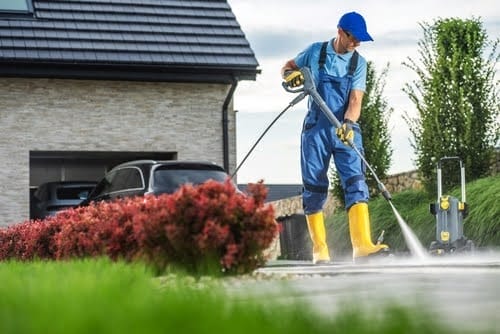 A man in blue overalls and yellow boots cleans a driveway with a pressure washer, surrounded by lush greenery and vibrant flowers on a bright afternoon.